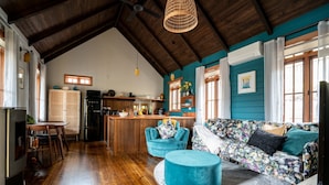 A spacious living area with a wooden ceiling, a kitchen in the background, and a teal-colored sofa with floral cushions. - Eliza's Bright (Bright)
