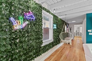 Una habitación con un muro cubierto de hojas verdes, un letrero de neón, un sillón colgante y una ventana con persianas.