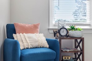 A blue armchair with a white and pink cushion, a small wooden side table with a clock and a potted plant, and a window with blinds. - Downtown Raleigh | Sleeps 15 | Game Yard & Parking (Raleigh)