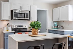 Cocina moderna con gabinetes blancos, electrodomésticos de acero inoxidable y una isla central con una cesta tejida que contiene plantas verdes.