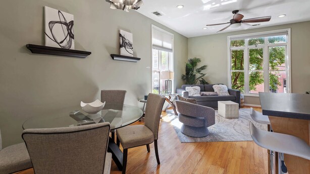 A modern living room with a glass dining table, comfortable seating, a ceiling fan, and large windows. - Warehouse District Modern Condo w/ Private Garage (Raleigh)