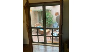 Une porte-fenêtre coulissante donne sur un patio extérieur doté d’un sol en briques, d’une chaise en osier et d’une chaise en plastique bleue.