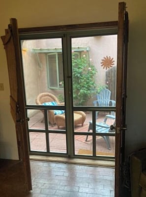 Eine Glastür öffnet sich zu einer Außenpatio-Terrasse mit Backsteinpflaster, einem Korbstuhl und einem blauen Plastikstuhl.