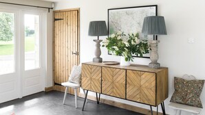A room with a wooden door, a white bench, a cabinet with geometric patterns, a vase with flowers, and two lamps.