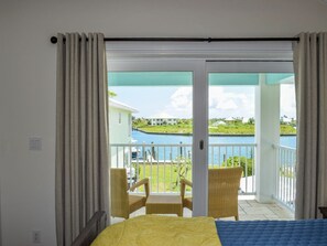 Quarto com cama, varanda com cadeiras de vime e vista para a água e edifícios.