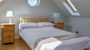 Ein Schlafzimmer mit schrägem Dach, einem Oberlicht, einem Bett mit weißer Bettwäsche, zwei Nachttischen mit Lampen und einem Nachttisch aus Holz mit Schubladen.