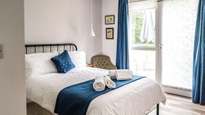 A neatly made bed with a blue blanket and two rolled towels, a chair, a small table, and a window with curtains. - 5 bedroom accommodation in Chilcompton (Chilcompton)