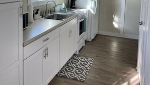 Una cocina moderna con armarios blancos, un lavamanos de acero inoxidable, un horno empotrado y un refrigerador. Hay estantes de madera sobre el lavamanos y una alfombra estampada en el suelo.