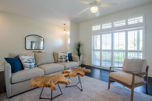 A living room with a sofa, a wooden coffee table, a chair, a ceiling fan, and large windows. - Beach House ! Sunset Decks ! Walk to Beach & John's Pass Boardwalk (Treasure Island)