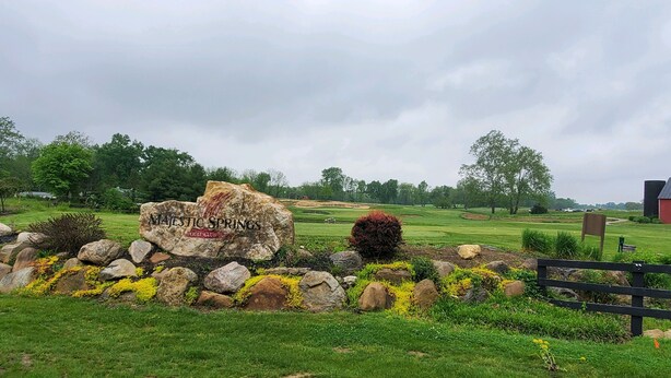 Un campo da golf con un grande cartello roccioso che riporta "Majestic Springs Golf Club", circondato da rocce e fiori gialli.