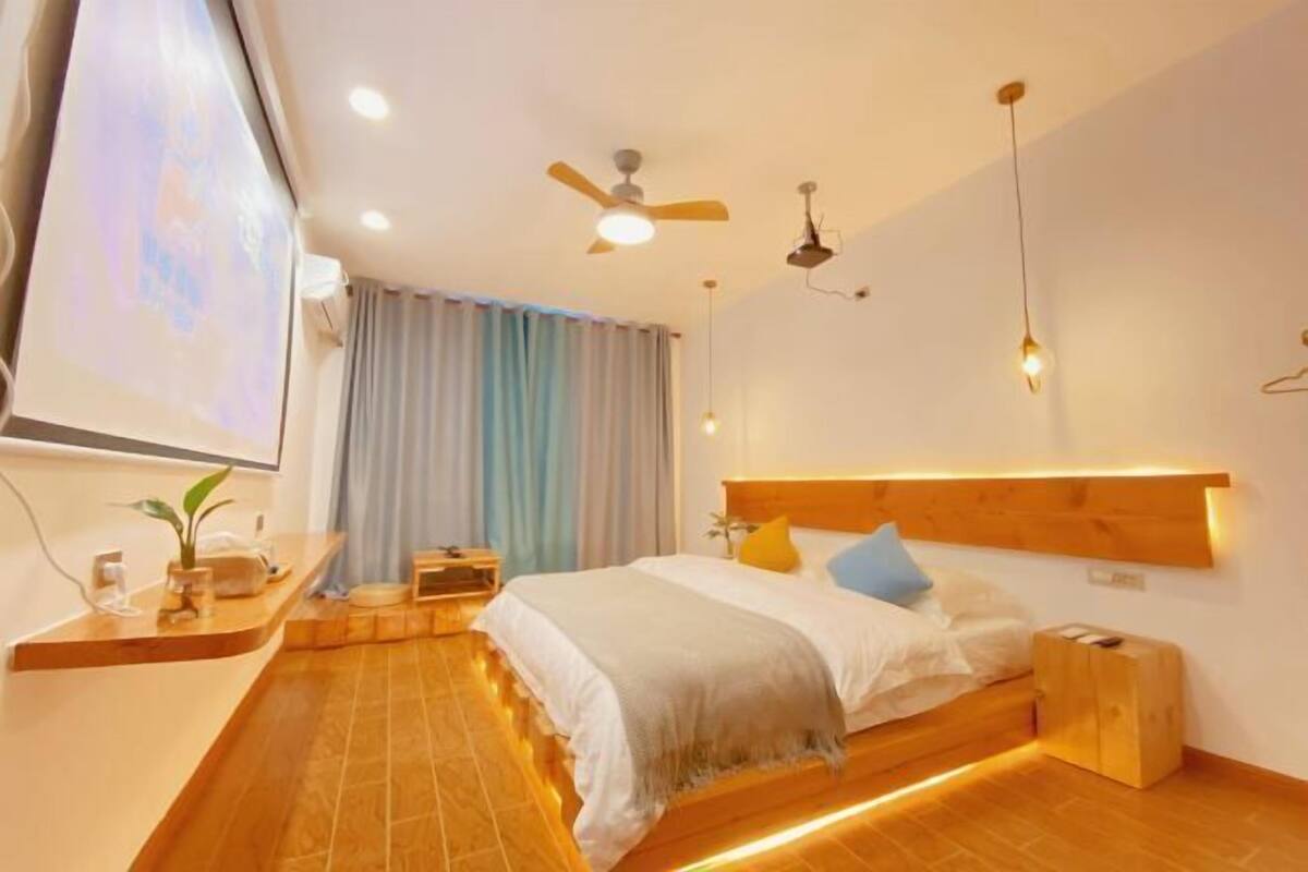 A modern bedroom with a wooden headboard, a bed with white bedding, a bedside table, a ceiling fan, and a wall-mounted projector.