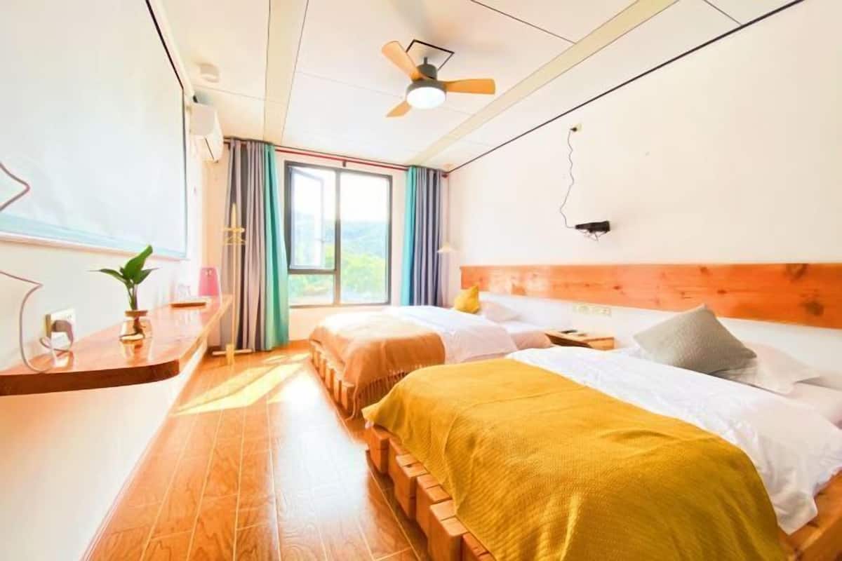 A room with two beds, a wooden headboard, a ceiling fan, a window with curtains, and a small table with a plant.