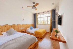 A hotel room with two beds, a ceiling fan, a window with curtains, and a wooden bedside table with a plant.