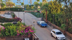 A street with a white car parked on the side, a blue car in the middle, and a playground with a slide and climbing structures.