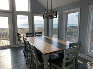 A dining area with a large wooden table and six chairs, large windows with ocean views, and a hanging light fixture.
