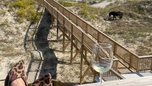 Ein hölzerner Steg führt zum Strand, auf einem Tisch steht ein Glas Wei ßwein und ein Paar leopardenmusterte Sandalen.