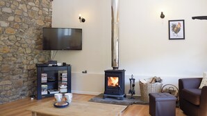 A cozy living room with a stone wall, a flat-screen TV, a wood-burning stove, a bookshelf, and a comfortable seating area. - A charming countryside cottage break. (Cwmdu, Near Llandeilo)