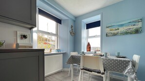 A dining area with a table set for four, white chairs, and a view of the outdoors through large windows. - 2 bedroom accommodation in Portmahomack (Portmahomack)