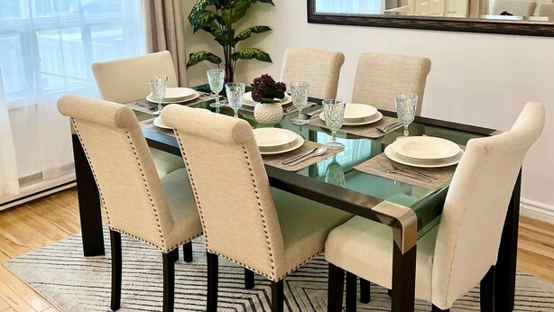 Un coin repas avec une table à plateau en verre, des chaises recouvertes d’un tissu beige et un lustre suspendu.