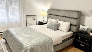 Une chambre à coucher moderne avec un lit recouvert d’un tissu gris, une table de chevet avec une lampe et une citation encadrée accrochée au mur.