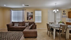 A living room with a brown sofa, a dining table set for four, and a kitchen area in the background. - This house is a 5 bedroom(s), 3 bathrooms, located in Eastvale , CA. (Eastvale)