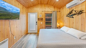 Een slaapkamer in een houten blokhut met een bed, een airconditioner, een deur en uitzicht op een bebost landschap.