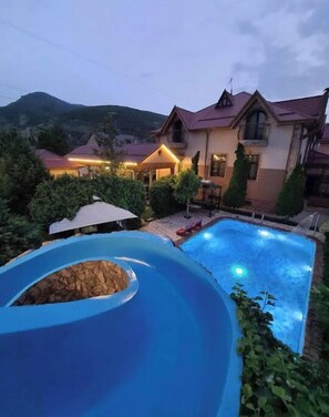 A swimming pool with a slide, surrounded by a landscaped garden and a large house with a sloped roof. - Villa "Story" in Chortoq, Tashkent (Тошкент)