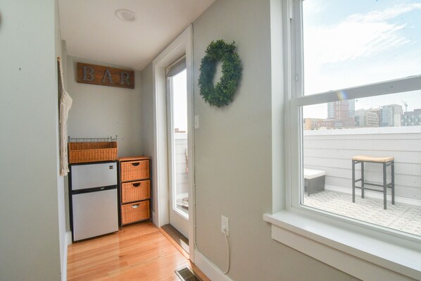 A small kitchen with a refrigerator, wooden cabinets, a window with a view of buildings, and a 'BAR' sign on the wall. - Minutes to Downtown & Waterfronts with rooftop deck! (Baltimore)