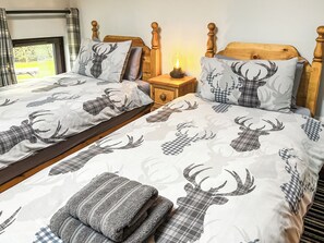 A double bed with a plaid deer print bedspread and matching pillows, a bedside table with a lamp, and a window with curtains.