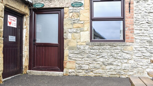 Ein Steinhaus mit einer dunkelbraunen Tür und einem Fenster. Schilder über der Tür zeigen 'Lathkill Suite' und 'Brennan Suite'. Ein Schild an der Tür verkündet: 'Frische Eier zum Verkauf'.