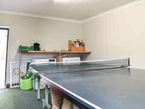Una mesa de ping pong en una habitación con un radiador, una cesta verde y una estantería con diversos objetos.