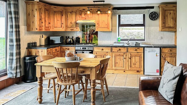 A kitchen with wooden cabinets, a dining table with chairs, a refrigerator, and a window with a view of greenery.