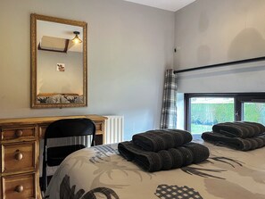 Un dormitorio con cama, escritorio, silla, espejo grande y ventana con vista a follaje.