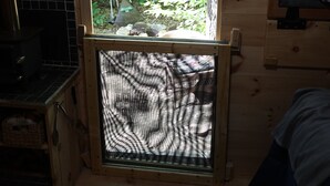Une petite cabane en bois avec une fenêtre donnant sur une forêt. À l’intérieur, on trouve une étagère en bois avec des paniers et un petit poêle.