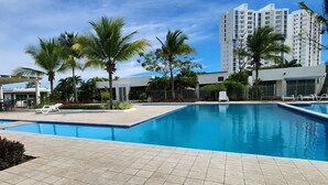 A swimming pool with lounge chairs and palm trees, surrounded by a modern building and a tall residential building in the background.