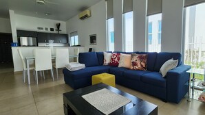 A modern living room with a blue sofa, a glass coffee table, and a yellow ottoman.