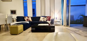Ein modernes Wohnzimmer mit einem dunkelblauen Sofa, einem gelben Hocker und einem schwarzen Couchtisch. Es gibt einen Glastisch mit einer Lampe und einer kleinen Pflanze. Das Zimmer hat große Fenster mit blauen Vorhängen und Blick ins Freie.