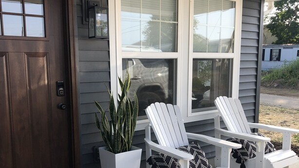 Un porche con una puerta marrón, dos sillas Adirondack blancas, una planta en maceta y un banco blanco.