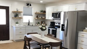 Una cocina moderna con armarios blancos, un refrigerador de acero inoxidable y un comedor con mesa y sillas de madera.
