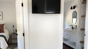 Habitación de hotel moderna con cama, televisor montado en una pared blanca, baño con lavamanos y espejo, y suelo de madera.