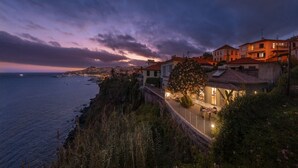 Un pueblo costero al anochecer, con edificios que dan al mar, un sendero pavimentado y un árbol con flores rosadas.