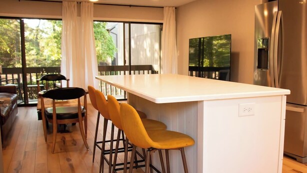 A modern kitchen with a white island, yellow bar stools, stainless steel appliances, and large windows with curtains. - The Duffer's Drop - 2BR/2BA Condo in Pinehurst, NC (Pinehurst)