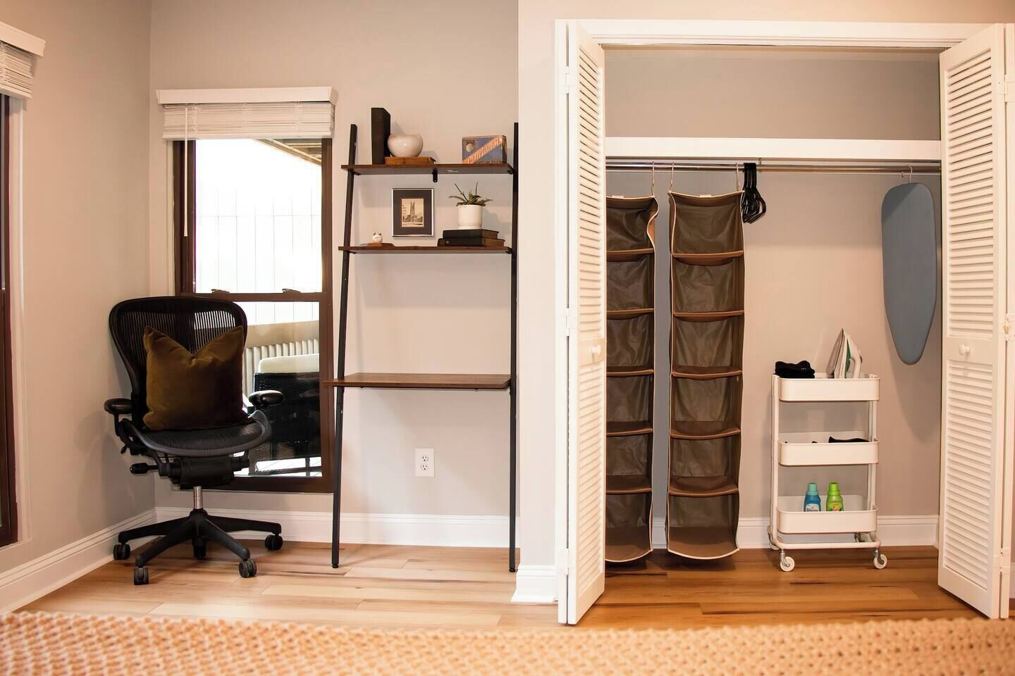 Una habitación con una silla de oficina negra, un estante blanco con libros y una planta, un armario blanco con espacio para colgar ropa, y un estante blanco con botellas.
