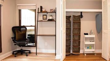 Una habitación con una silla de oficina negra, un estante blanco con libros y una planta, un armario blanco con espacio para colgar ropa, y un estante blanco con botellas.