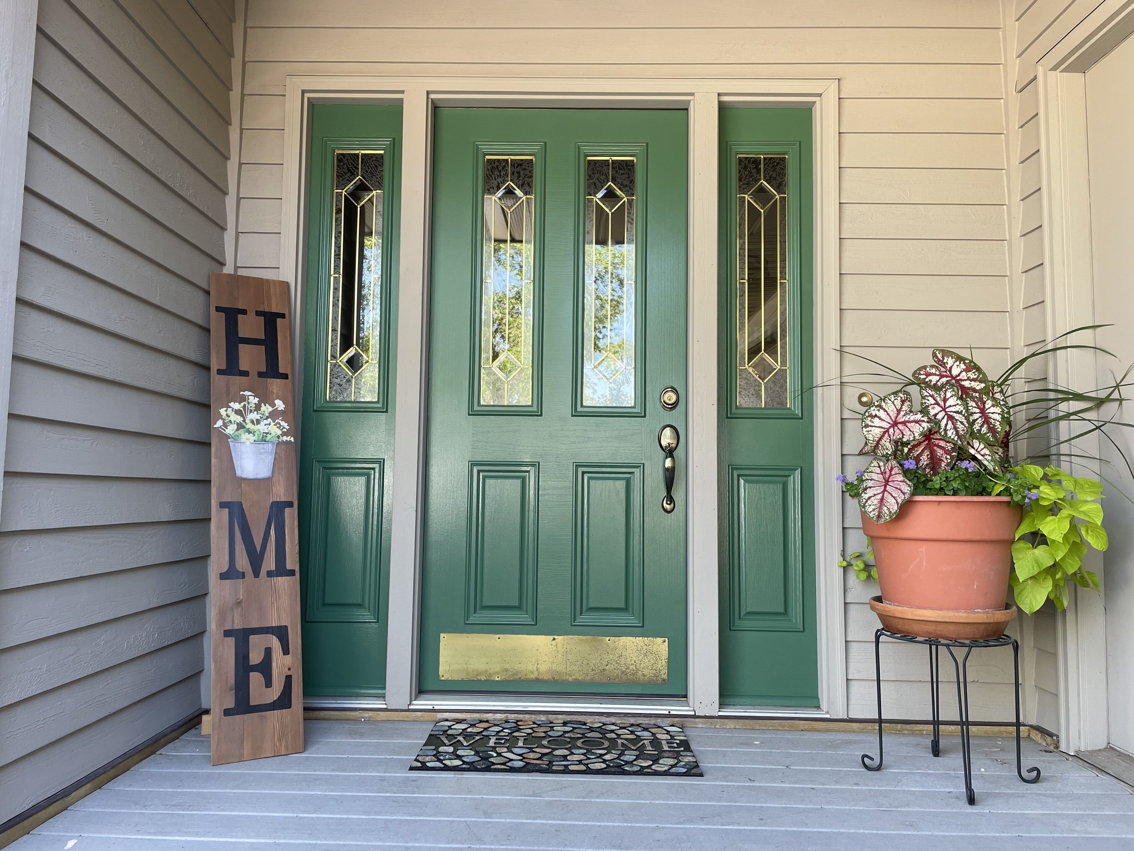 A green door with a 'WELCOME' sign, a doormat, and a potted plant on a stand.