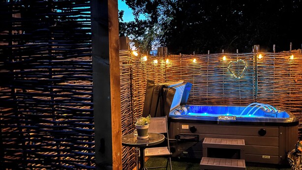 Zona de hidromasaje al aire libre con valla de madera, luces colgantes y una mesa con sillas.
