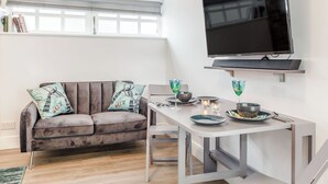 Una sala de estar moderna con un sofá gris, una mesa de comedor con dos sillas, un televisor de pantalla plana montado en la pared y una ventana con persianas.