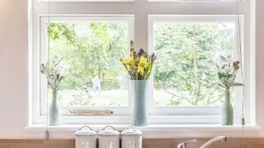 Una cocina con vista al jardín por la ventana, un florero con flores y tres recipientes metálicos sobre la encimera.