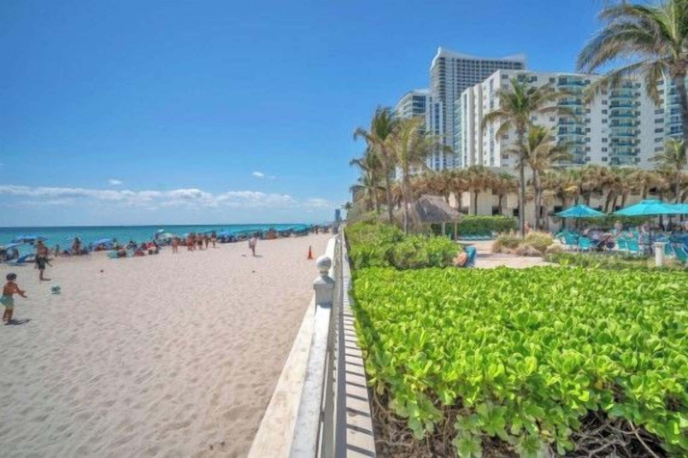 A beach with a pool area, palm trees, and high-rise buildings in the background.