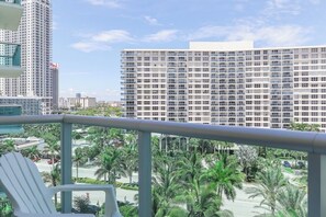 Un balcon avec une chaise blanche, donnant sur un paysage urbain avec de hauts immeubles et des palmiers.